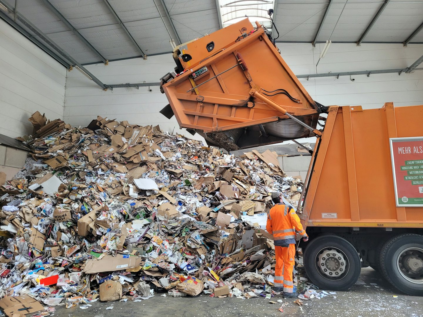 Trabajador observa cómo camión de basura naranja descarga cartón en planta de reciclaje.